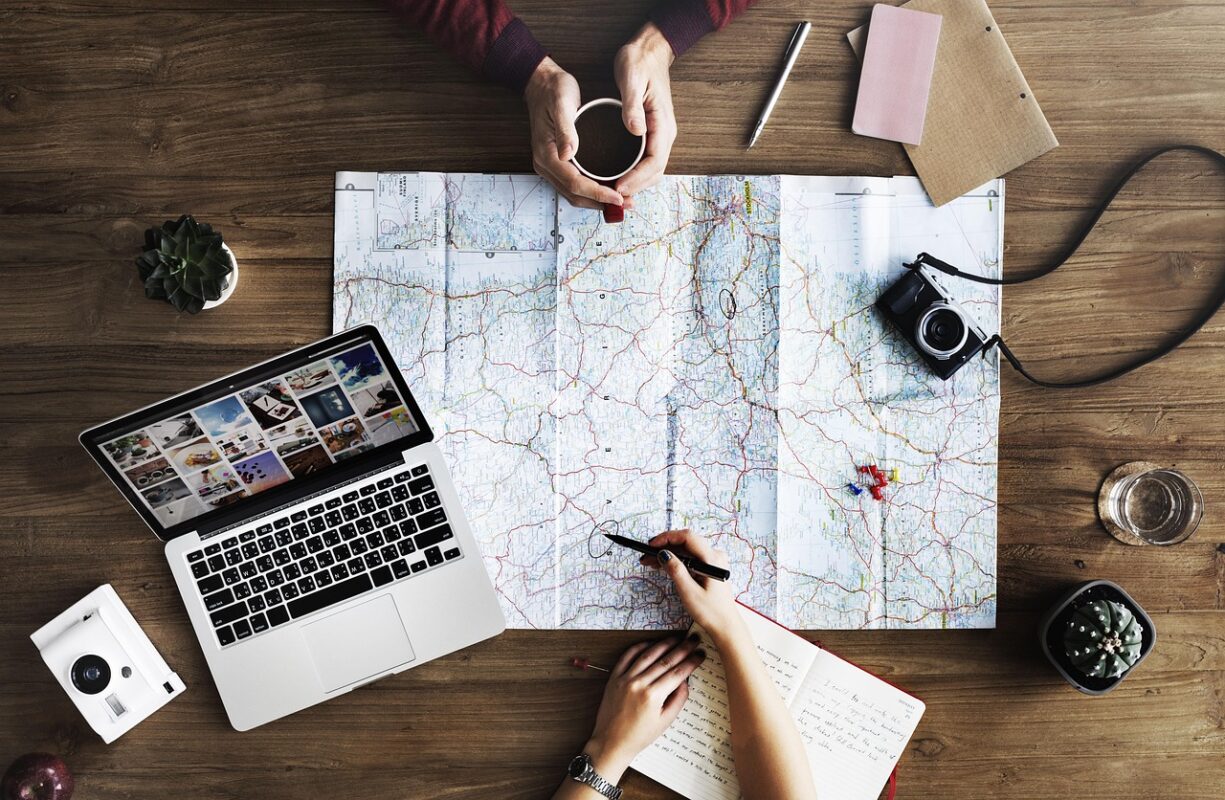 hands of two people sitting across from each other planning travel with laptop and one person showing routes on a paper map with a digital camera on the map.
