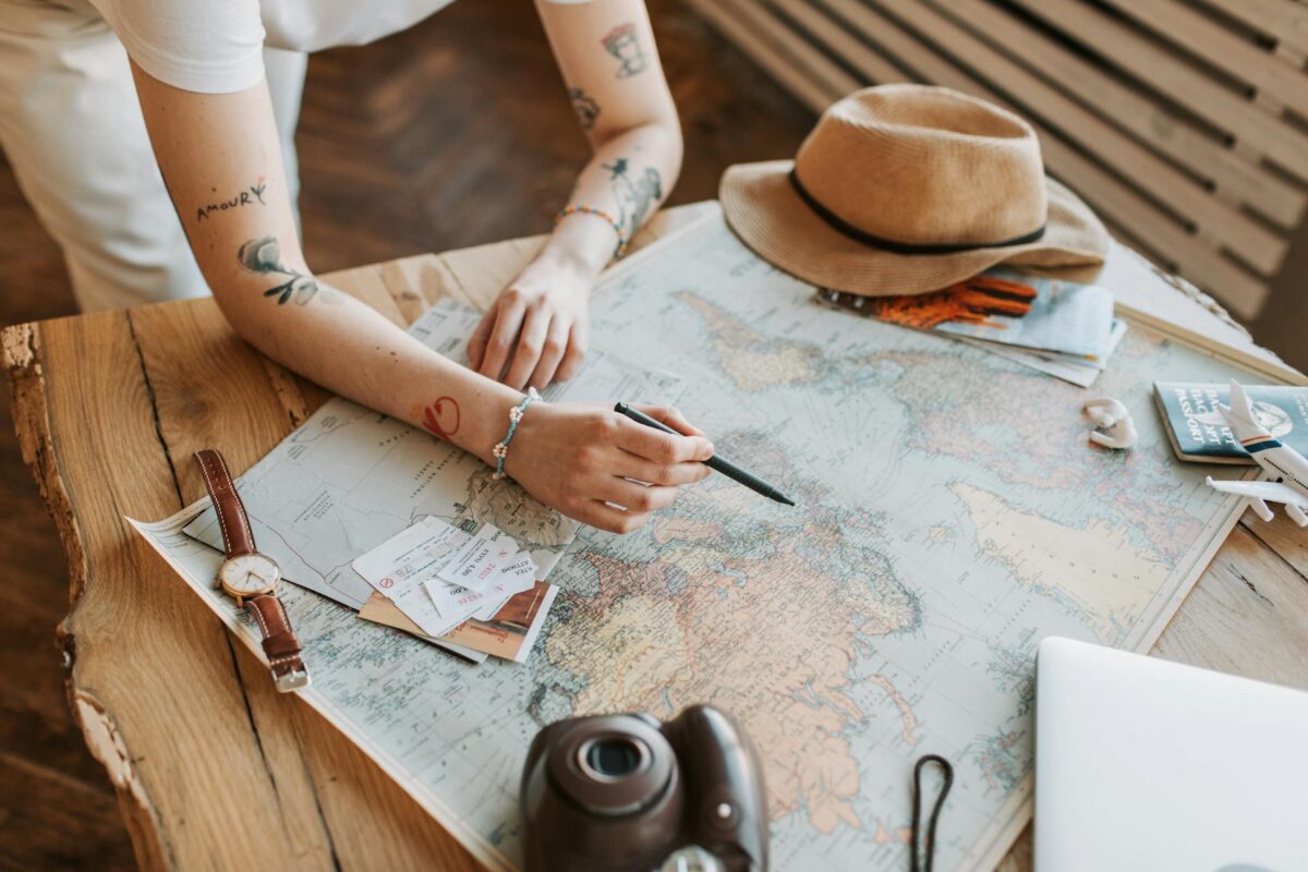 A woman checking a world map, holding a pen. A watch, hat, passport and polaroid camera is on the map