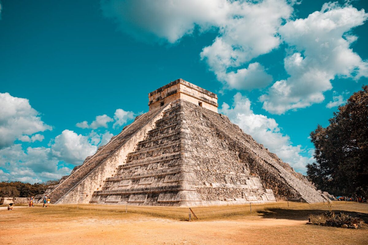A low-angle, wide shot of the Chichén Itzá pyramid in Mexico under a bright blue sky with fluffy white clouds. The pyramid, also known as El Castillo, is made of light-colored stone, with visible stairs climbing each of its four sides. The ground in the foreground is sandy and brown, with a few people walking in the distance to the left. A large, green tree is visible on the right side of the frame.