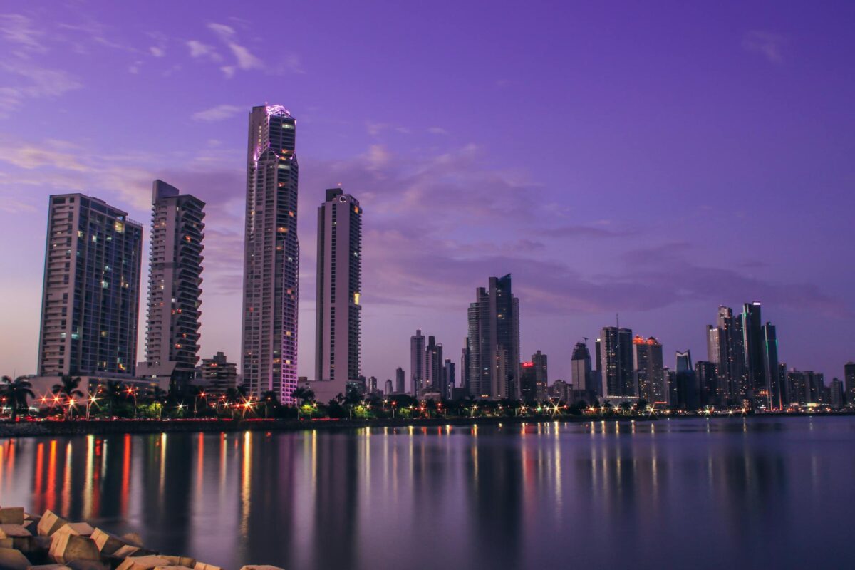 A panoramic, wide-angle shot of the Panama City skyline at sunset. Modern, high-rise buildings and skyscrapers are illuminated against a vibrant purple and pink sky, reflecting off the calm water in the foreground. The streetlights along the waterfront are on, creating streaks of light and adding to the city's lively atmosphere. The image showcases the modern and beautiful cityscape, highlighting the appeal of living in Panama City as an expat.