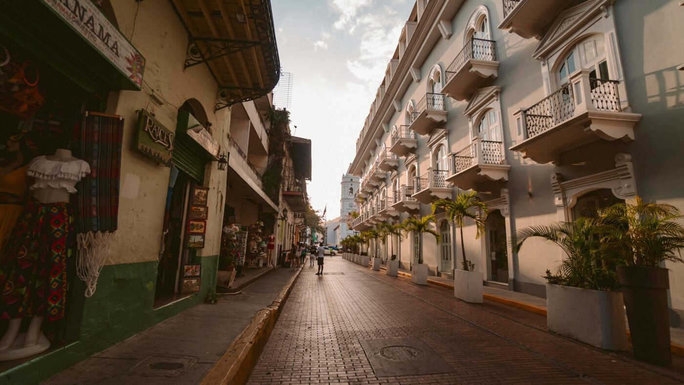 A street view in Casco Viejo, Panama City, with old colonial buildings lining both sides of a cobblestone street. The street is empty except for a few pedestrians in the distance, and the sun is shining down, illuminating the path. The buildings on the left are shops with awnings and mannequins, while the buildings on the right are residential with balconies and potted plants. The image captures the essence of living in Panama City as an expat, highlighting the unique mix of historical architecture and vibrant street life.