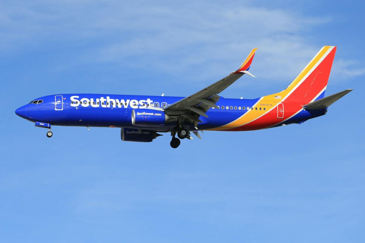 A full-side view of a Southwest Airlines plane flying low in the sky, with its landing gear down. The plane is painted in the airline's distinctive blue, red, and yellow livery, with "Southwest" written in large white letters on the fuselage. The image captures the plane in motion, a common sight for travelers familiar with the Southwest Airlines baggage policy.
