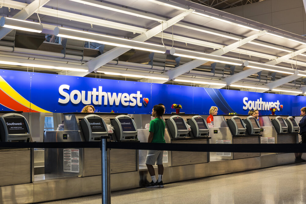 A wide, brightly lit shot inside an airport terminal, showing the Southwest Airlines check-in counters. Several numbered kiosks are lined up, each displaying the Southwest logo. Passengers are standing in line, and airline staff are visible behind the counters. The scene represents a typical airport check-in process, relevant to understanding the Southwest Airlines Baggage Policy before boarding a flight.