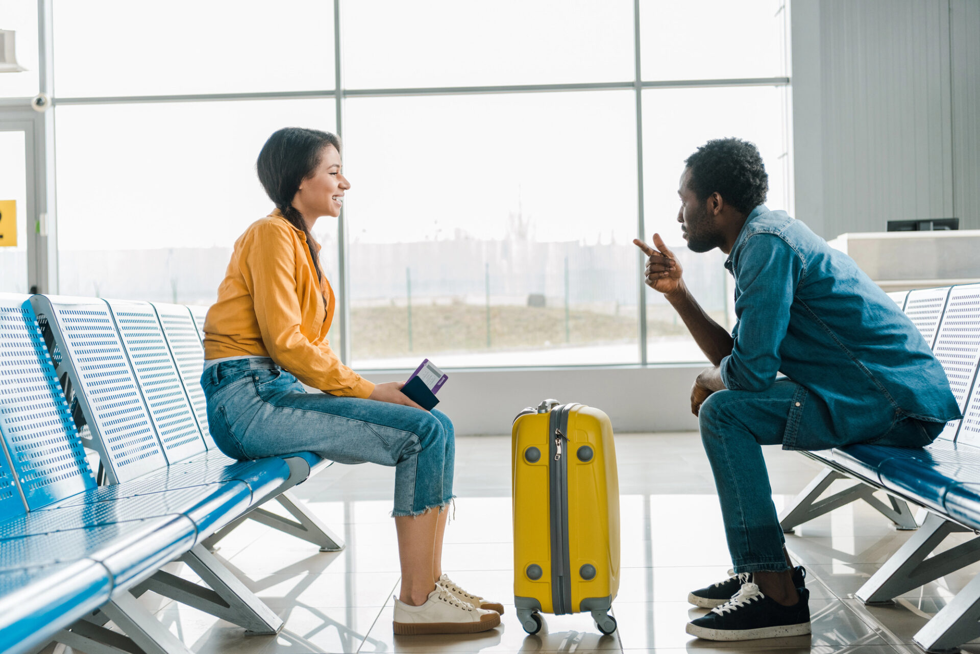 Engaging image showcasing two young adults seated in an airport terminal, highlighting a lively conversation. The woman, dressed in a vibrant orange top, smiles as she holds travel documents, while the man, in a blue denim shirt, gestures animatedly. A yellow suitcase adds a pop of color, creating a friendly atmosphere.